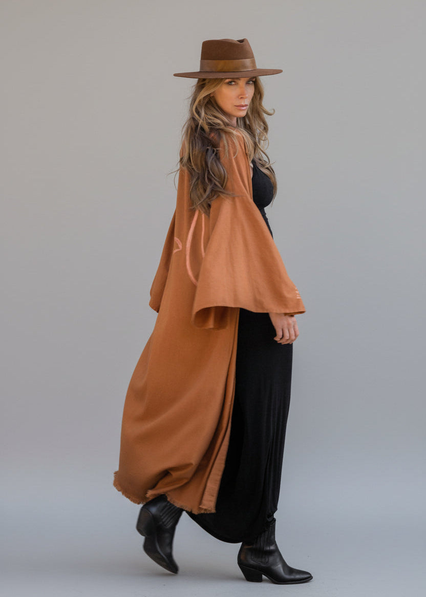 Woman wearing a hand-painted long kimono by House of Malibu styled with a hat over a tight black dress with boots